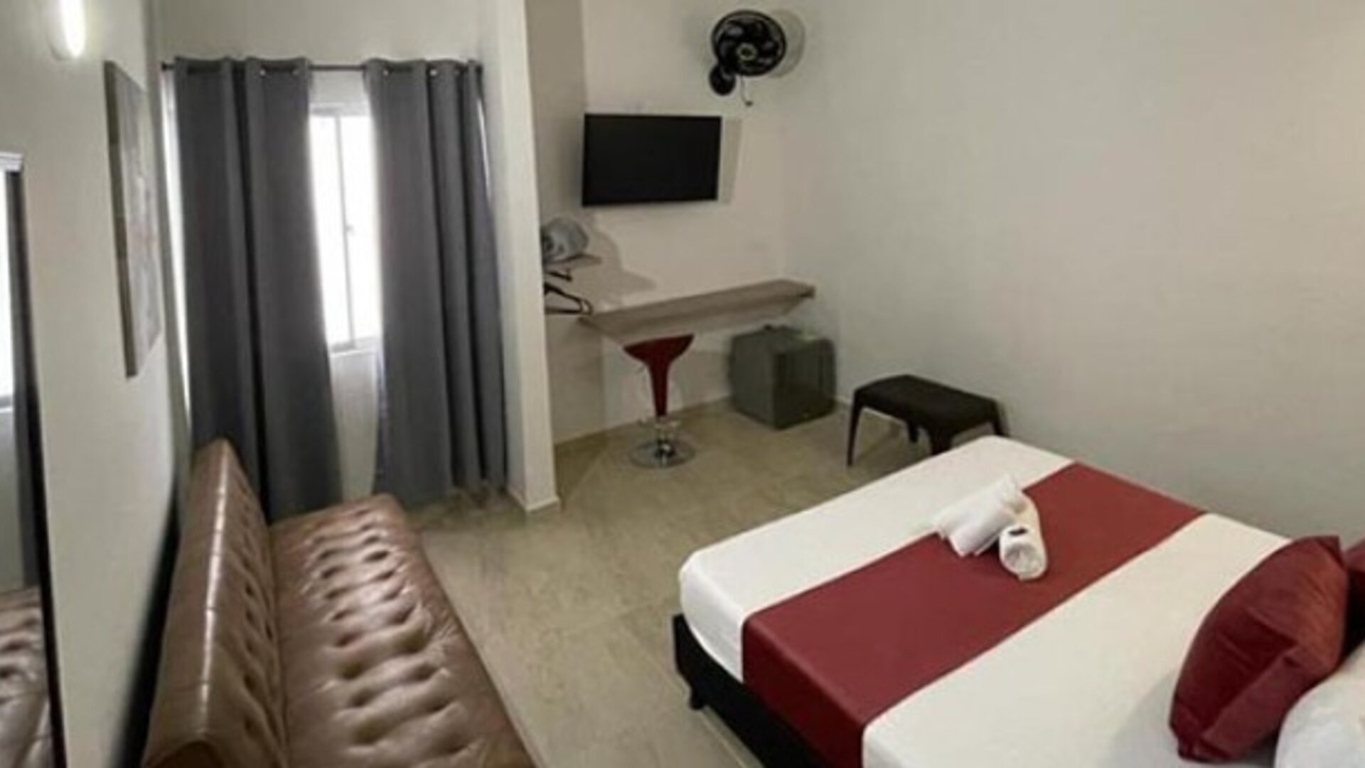 Guest room with double bed, red runner and cushions, brown sofa, wall-mounted television and curtained window at 12 Nights Hotel.