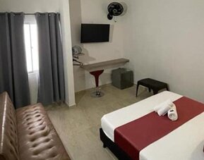 Guest room with double bed, red runner and cushions, brown sofa, wall-mounted television and curtained window at 12 Nights Hotel.