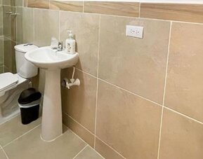 Bathroom with beige tiled walls, glass shower enclosure, pedestal sink, toilet and soap dispenser at 12 Nights Hotel.