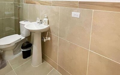 Bathroom with beige tiled walls, glass shower enclosure, pedestal sink, toilet and soap dispenser at 12 Nights Hotel.