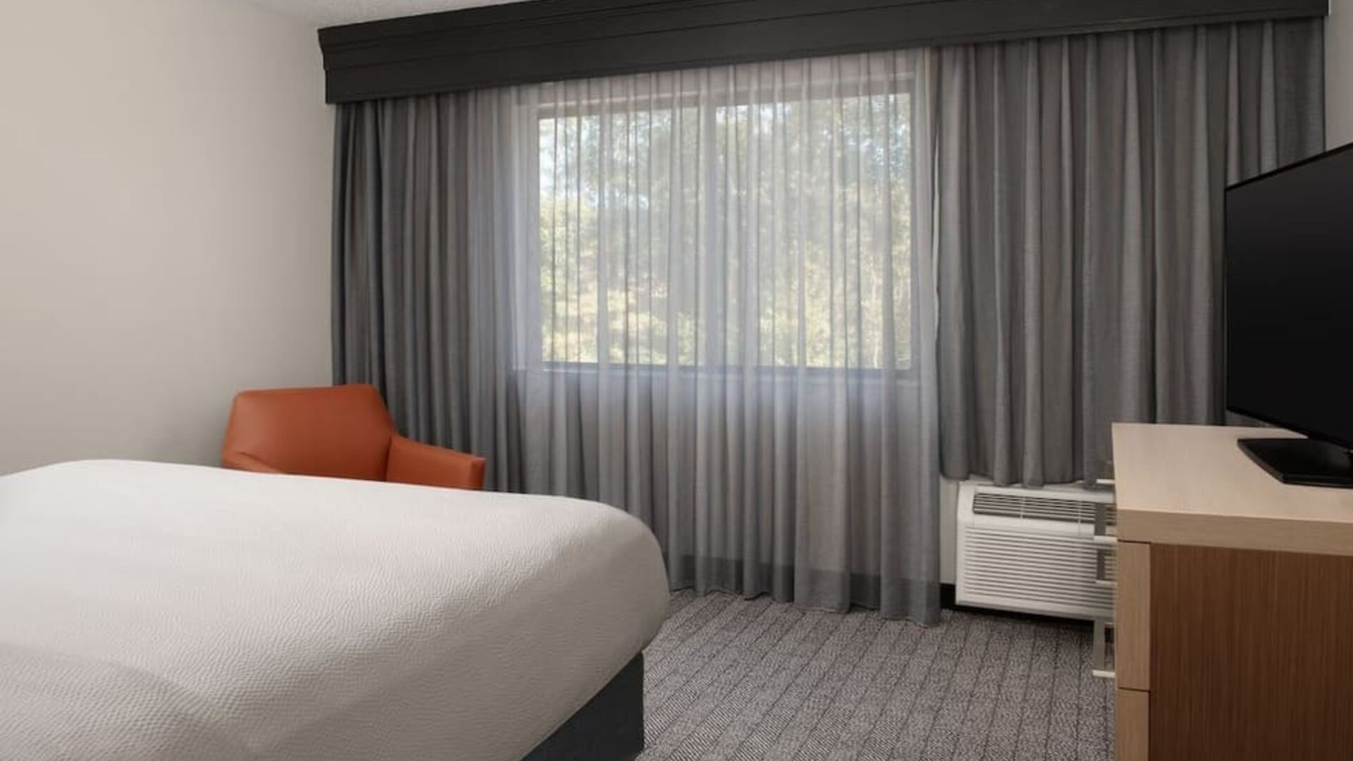 Guest room with white bed, orange accent chair, large curtained window, television and wooden desk at Courtyard By Marriott Dothan.