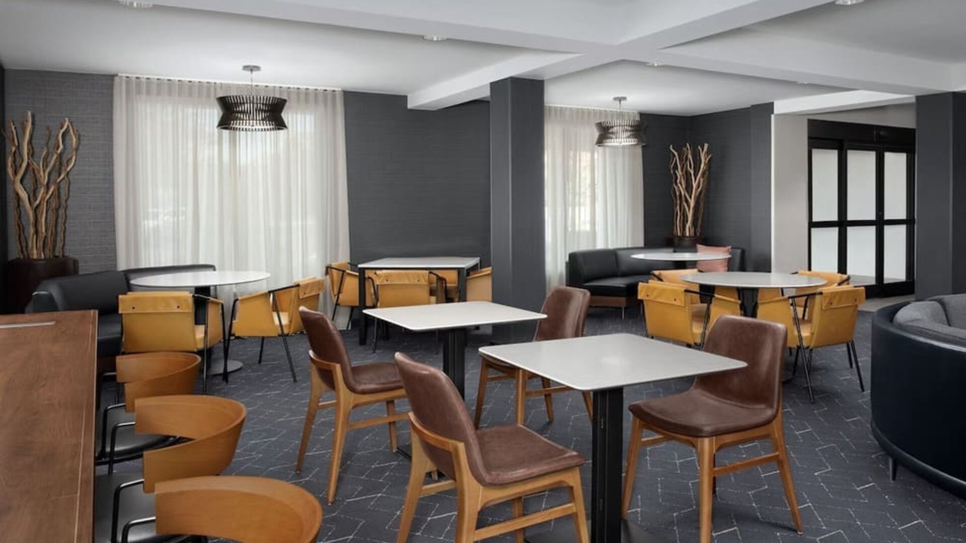Hotel lounge and dining area with tables, mustard-yellow chairs, brown armchairs, dark walls and large curtained windows at Courtyard By Marriott Dothan.