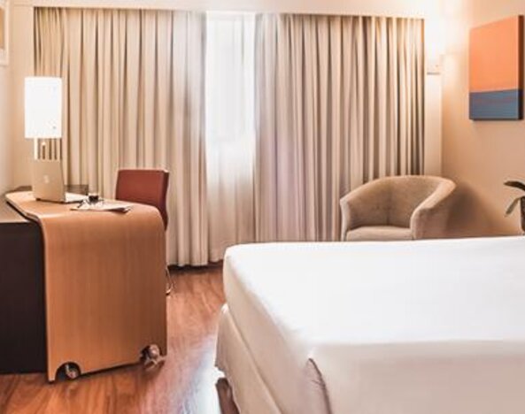 Guest room with king bed, orange accent cushions, wooden desk, armchair, television and floor-to-ceiling curtains at Novotel Lima San Isidro.