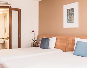 Twin guest room with white bedding, blue accent cushions, wooden headboards and an open ensuite bathroom at Novotel Lima San Isidro.