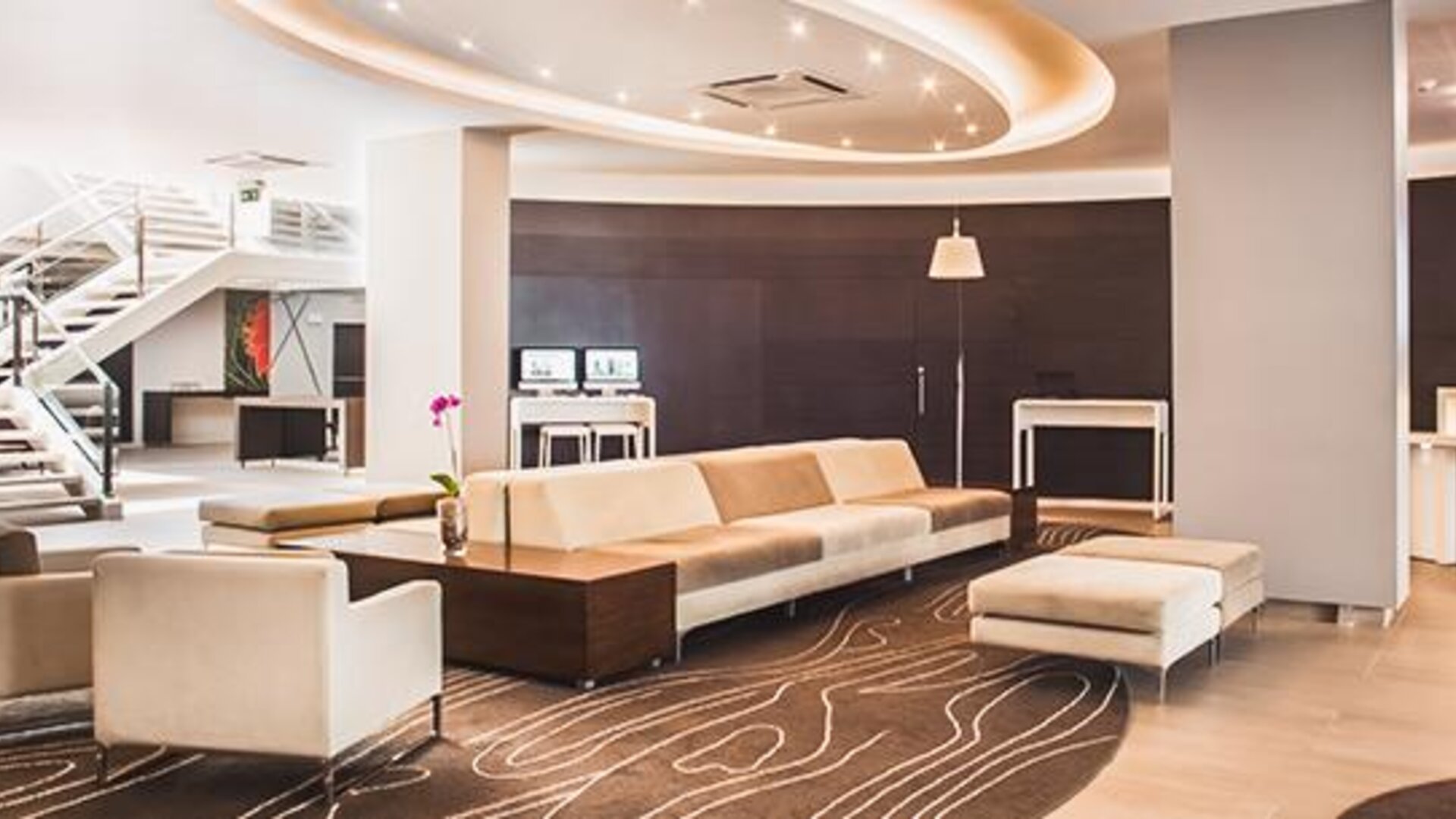 Hotel lounge with cream sectional sofas, patterned rug, staircase, decorative orchids and curved ceiling lighting at Novotel Lima San Isidro.