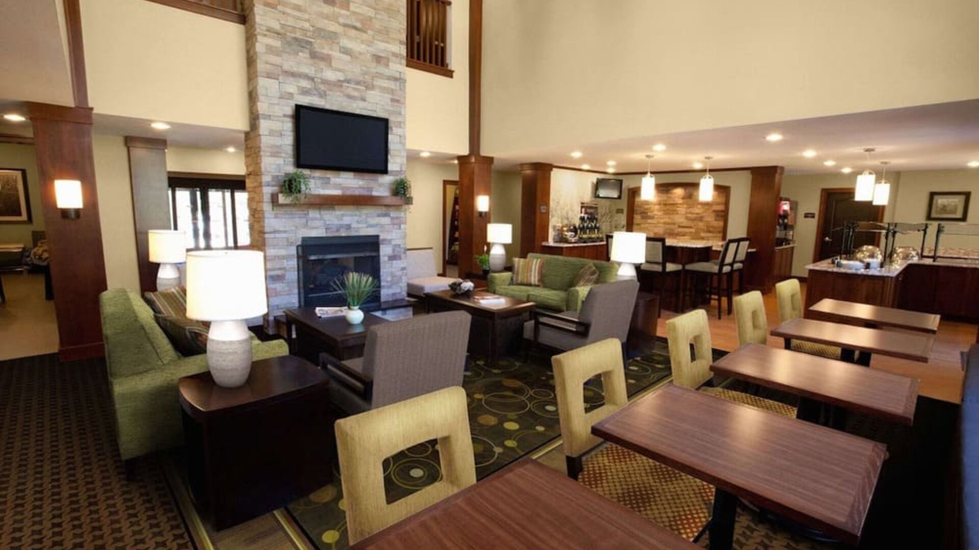 The hotel’s lounge is furnished with armchair and sofa seating around coffee tables, with a wall-mounted TV, and more tables and chairs nearby.