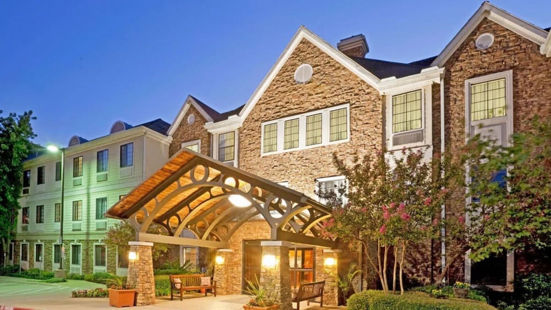 Sonesta ES Suites Dallas Las Colinas’ exterior has a covered entrance, benches, and greenery including potted plants, well-kept bushes, and trees.