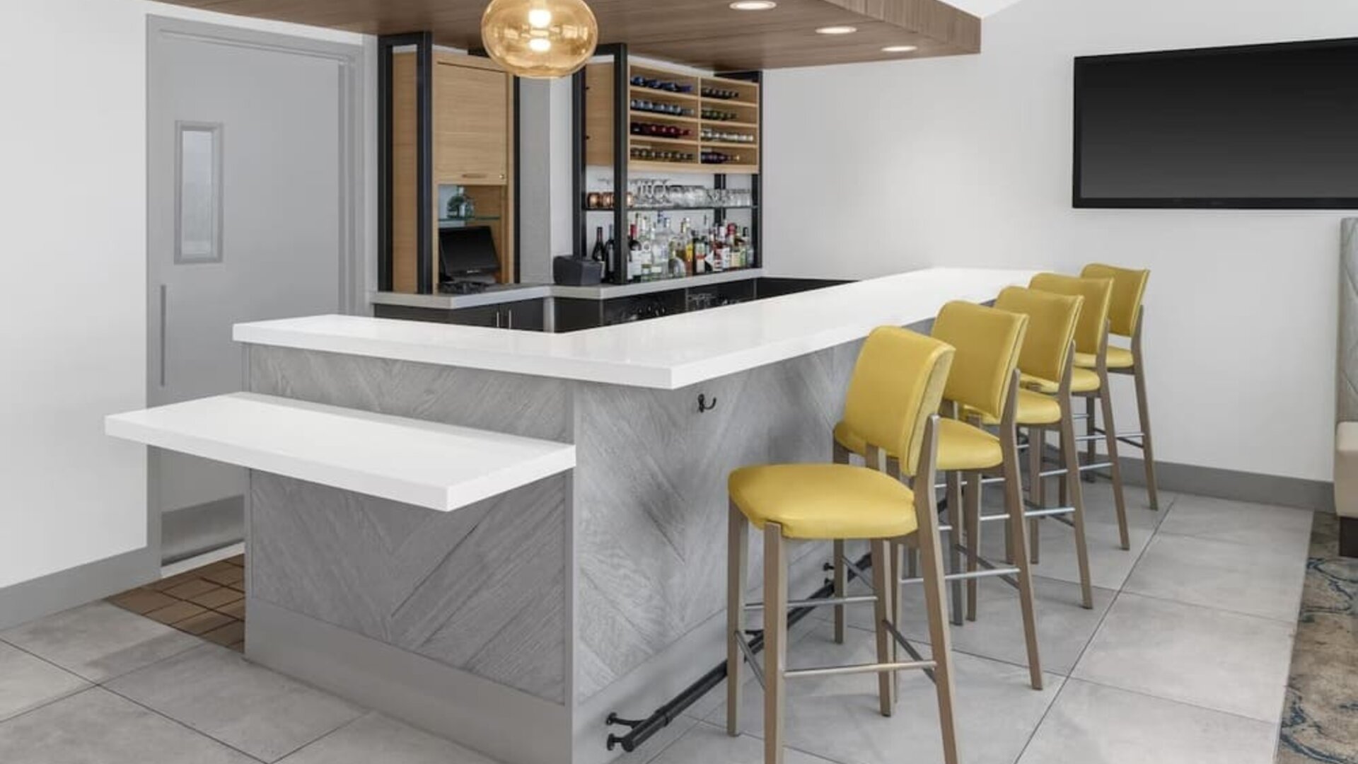 Hotel bar at Hilton Garden Inn Queens/JFK Airport with white counter, yellow stools, and bottle display.