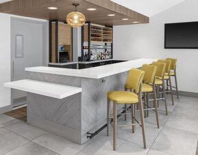 Hotel bar at Hilton Garden Inn Queens/JFK Airport with white counter, yellow stools, and bottle display.