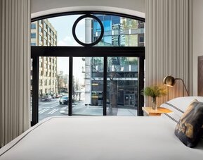 Bedroom with large window showing city street view, white bedding, and neutral decor at Kimpton Palladian Hotel.