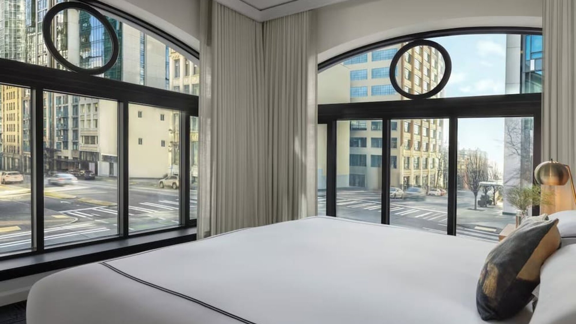 Corner bedroom with large curved windows, city street views, and white bedding at Kimpton Palladian Hotel.