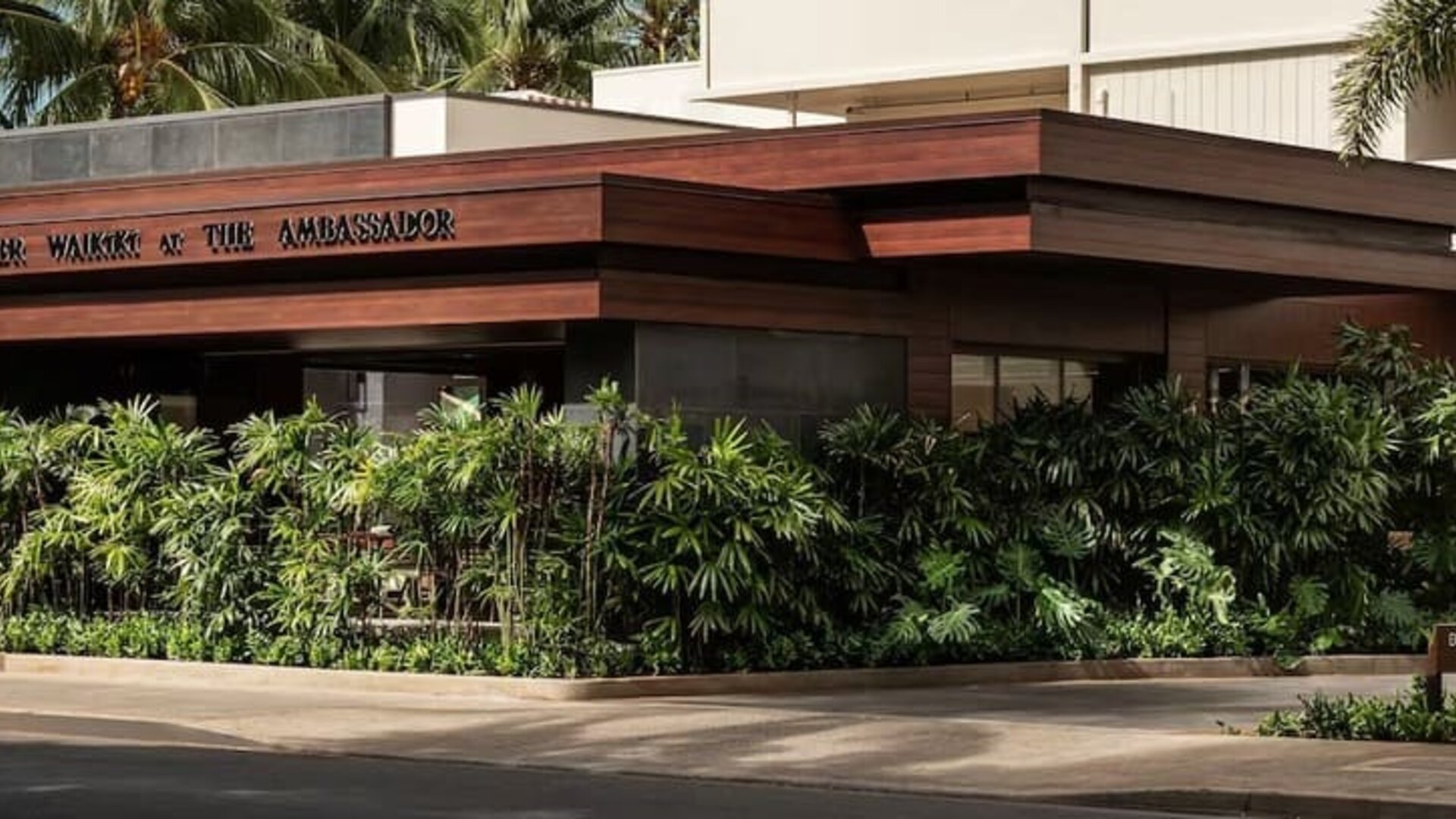 Exterior view of hotel entrance with tropical landscaping and signage at The Ambassador Hotel Of Waikiki.