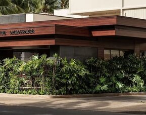 Exterior view of hotel entrance with tropical landscaping and signage at The Ambassador Hotel Of Waikiki.