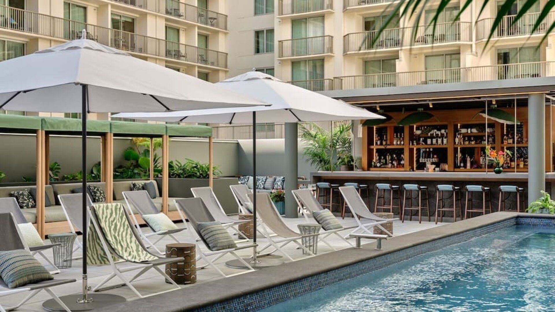 Outdoor pool with sun loungers, umbrellas, tiled deck, and mural wall surrounded by greenery at Romer House Waikiki.
