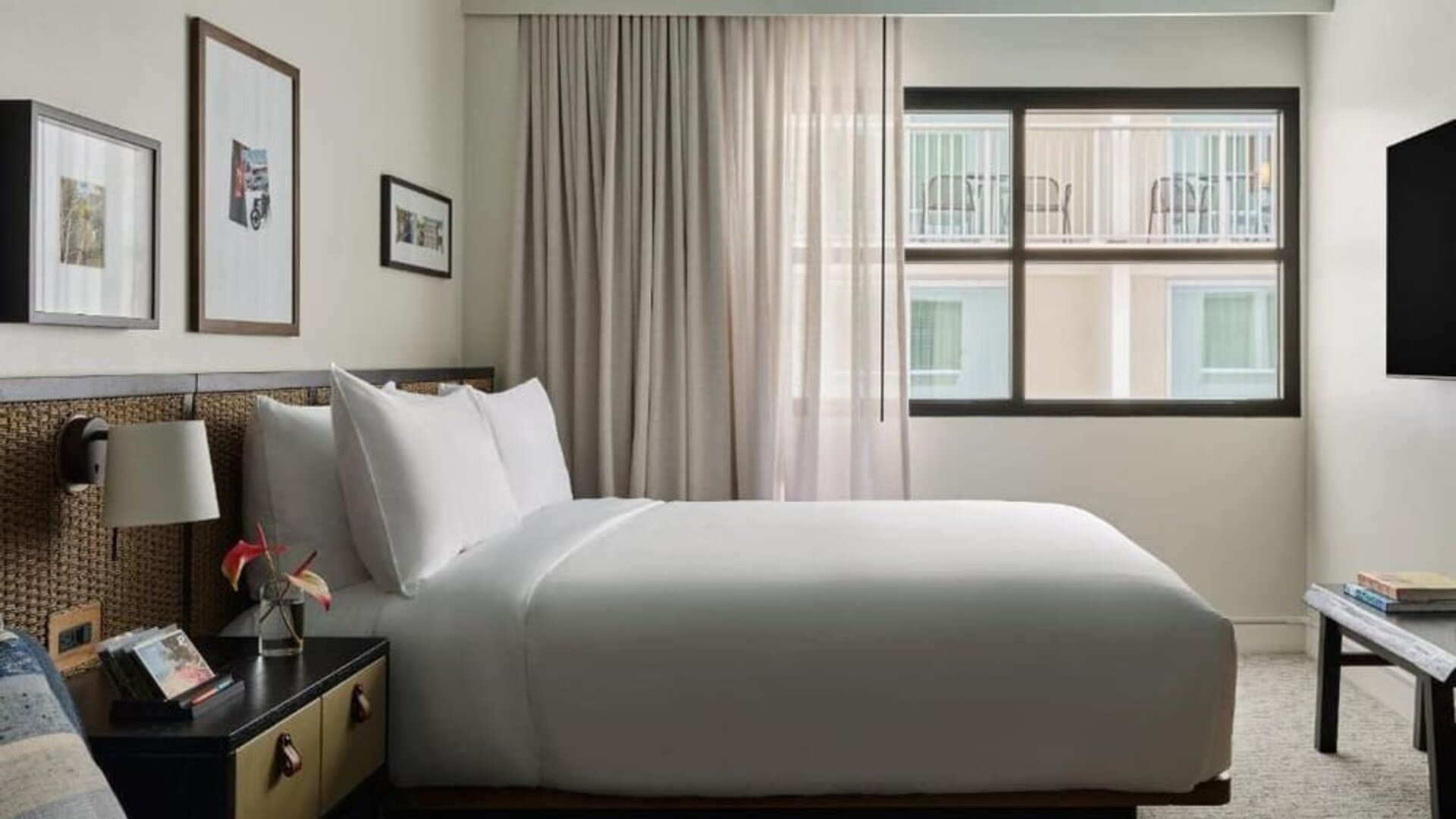 Queen bed with woven headboard beside window, soft curtains and framed wall art in a cozy layout at Romer House Waikiki.