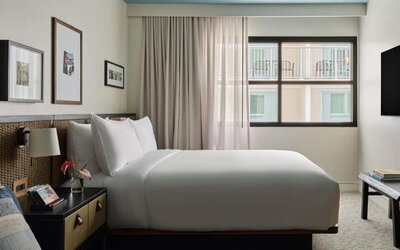 Queen bed with woven headboard beside window, soft curtains and framed wall art in a cozy layout at Romer House Waikiki.