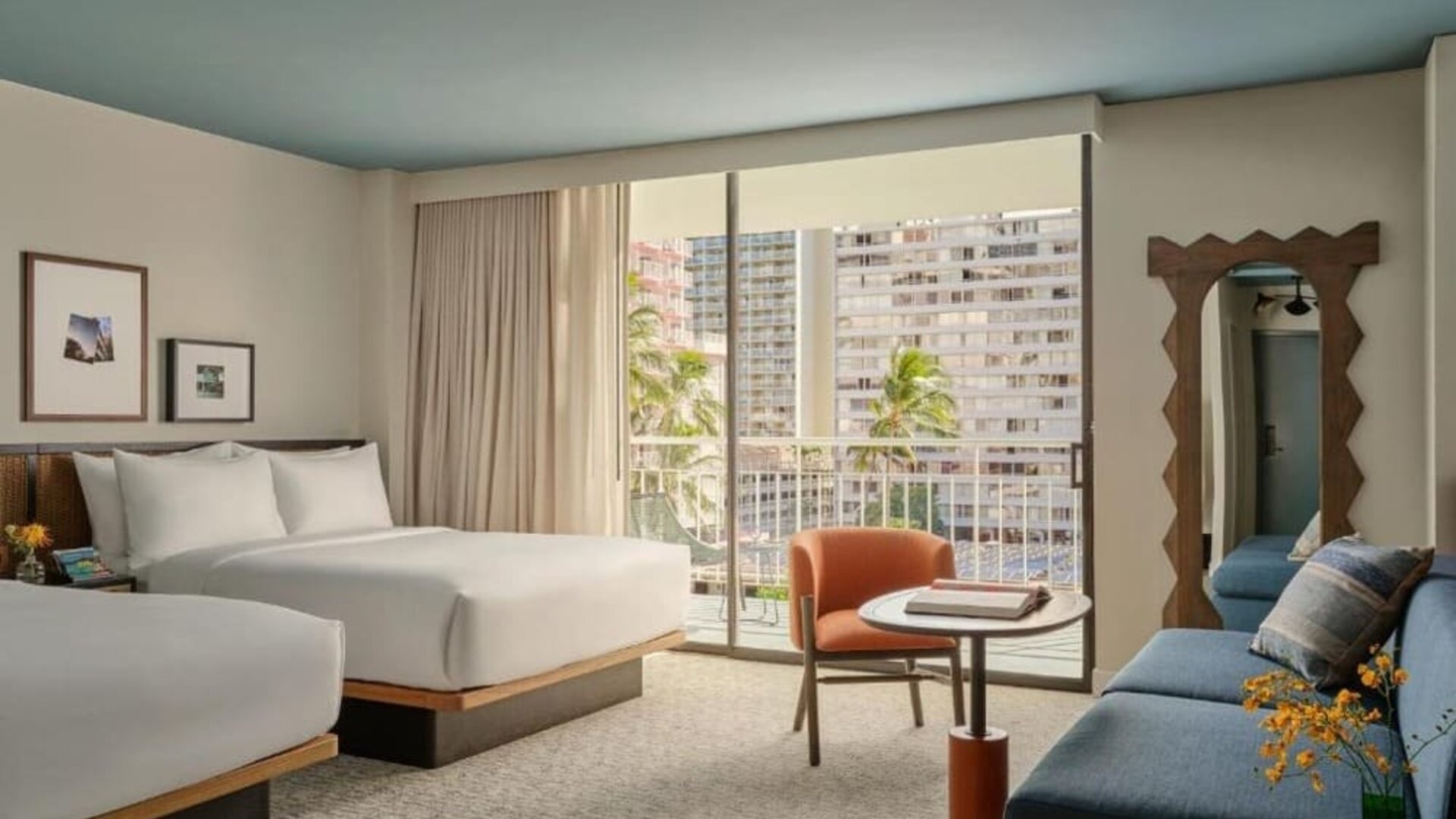 Guest room with beds near large window, balcony view of buildings, and seating area with chair and table at Romer House Waikiki.