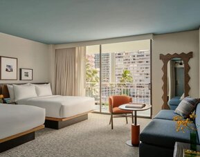 Guest room with beds near large window, balcony view of buildings, and seating area with chair and table at Romer House Waikiki.