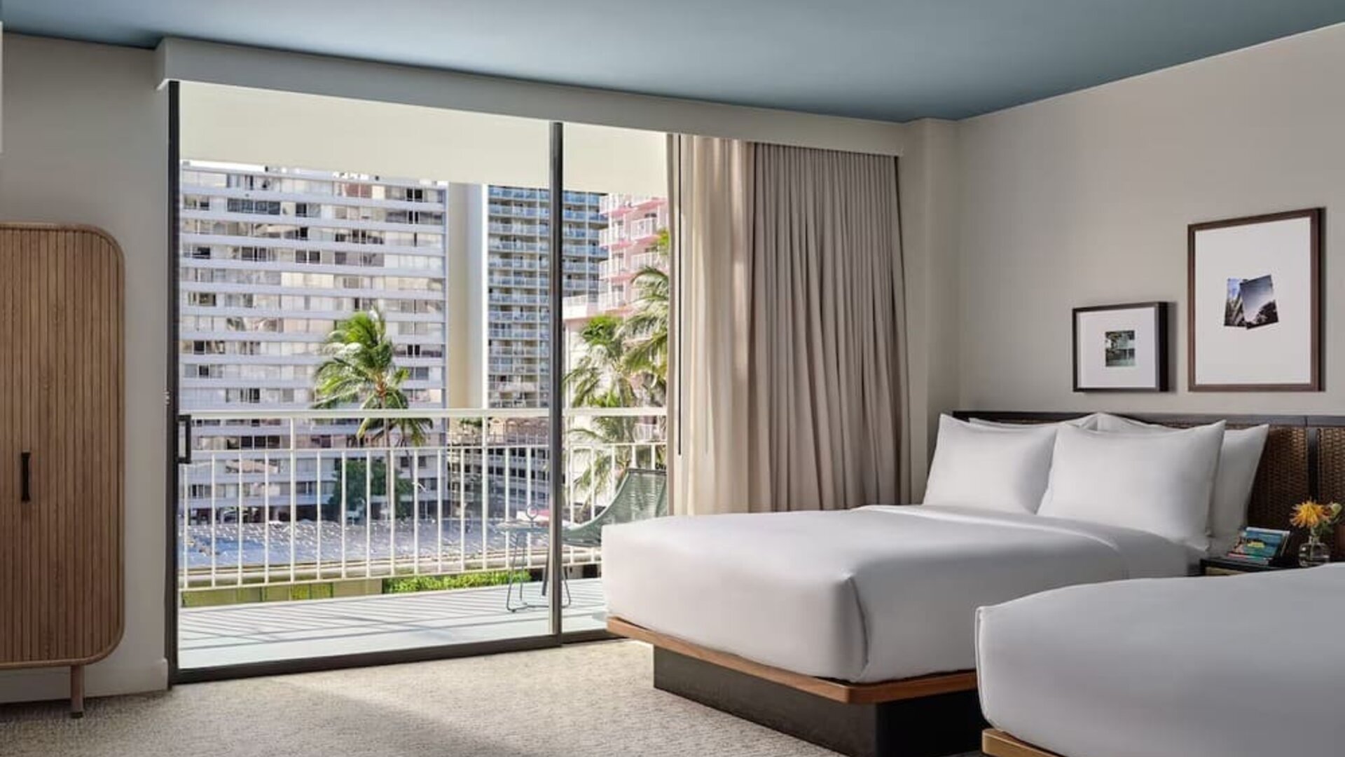 Two beds with white linens, balcony access, city view, and seating area with sofa and table at Romer House Waikiki.