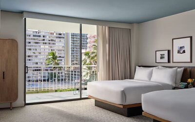 Two beds with white linens, balcony access, city view, and seating area with sofa and table at Romer House Waikiki.