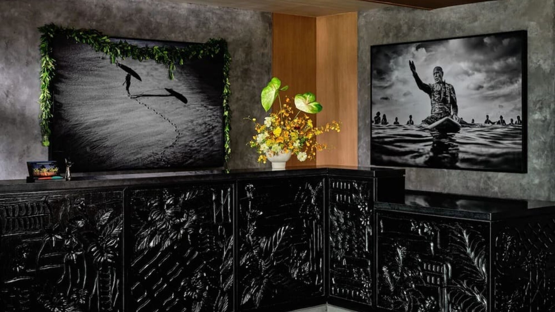 Reception desk with dark carved front panels, floral arrangement, and framed black-and-white artwork behind at Romer House Waikiki.