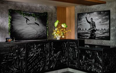 Reception desk with dark carved front panels, floral arrangement, and framed black-and-white artwork behind at Romer House Waikiki.