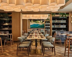 Restaurant dining space with long tables, cushioned chairs, wood accents, and decorative ceiling lighting at Romer House Waikiki.