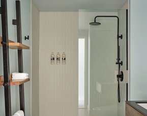 Bathroom with walk-in glass shower, black fixtures, white tile walls, and wooden shelving with towels at Romer House Waikiki.