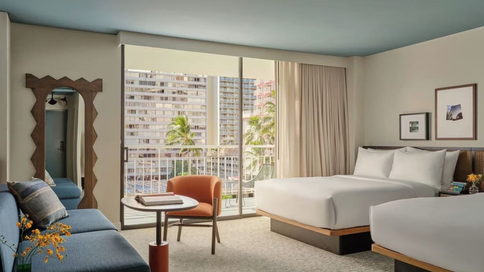 Two double beds with white linens, blue sofa, balcony access, and city view through sliding doors at Romer House Waikiki.