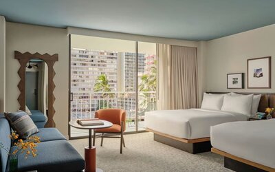 Two double beds with white linens, blue sofa, balcony access, and city view through sliding doors at Romer House Waikiki.