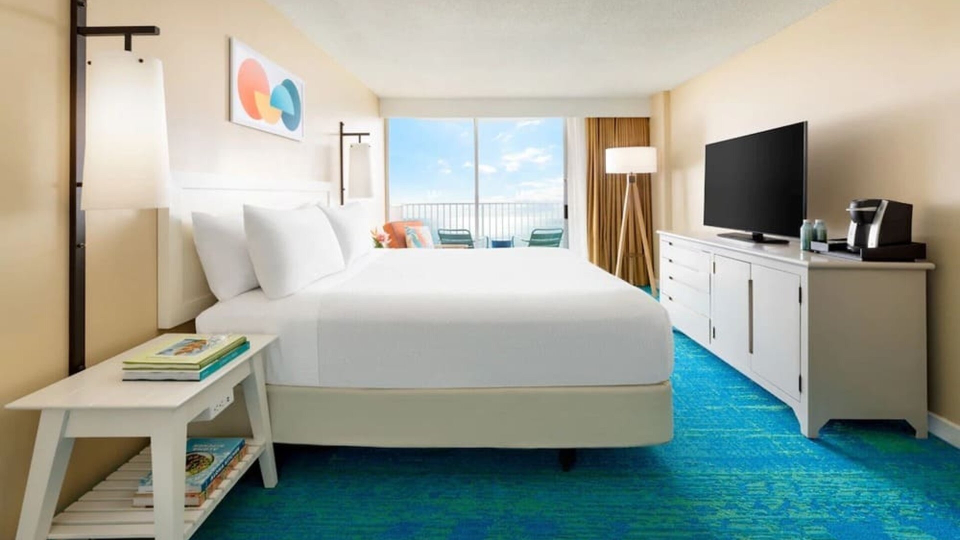 King bed facing balcony with ocean view, TV console, and blue patterned carpet at The Twin Fin Hotel.