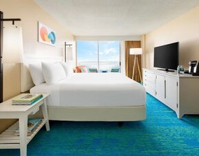 King bed facing balcony with ocean view, TV console, and blue patterned carpet at The Twin Fin Hotel.