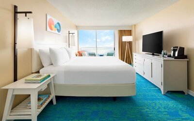 King bed facing balcony with ocean view, TV console, and blue patterned carpet at The Twin Fin Hotel.