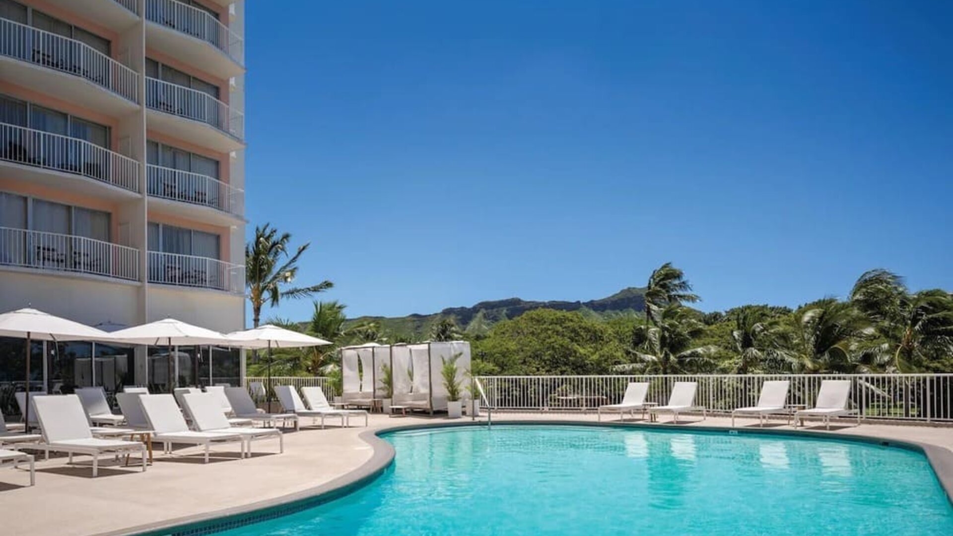 Outdoor pool with lounge chairs, umbrellas, and views of surrounding greenery at Park Shore Waikiki.