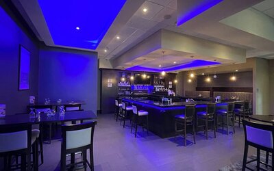 Hotel bar with high stools, ambient blue lighting, and dining tables at Holiday Inn & Suites Farmington Hills - Detroit NW By IHG.
