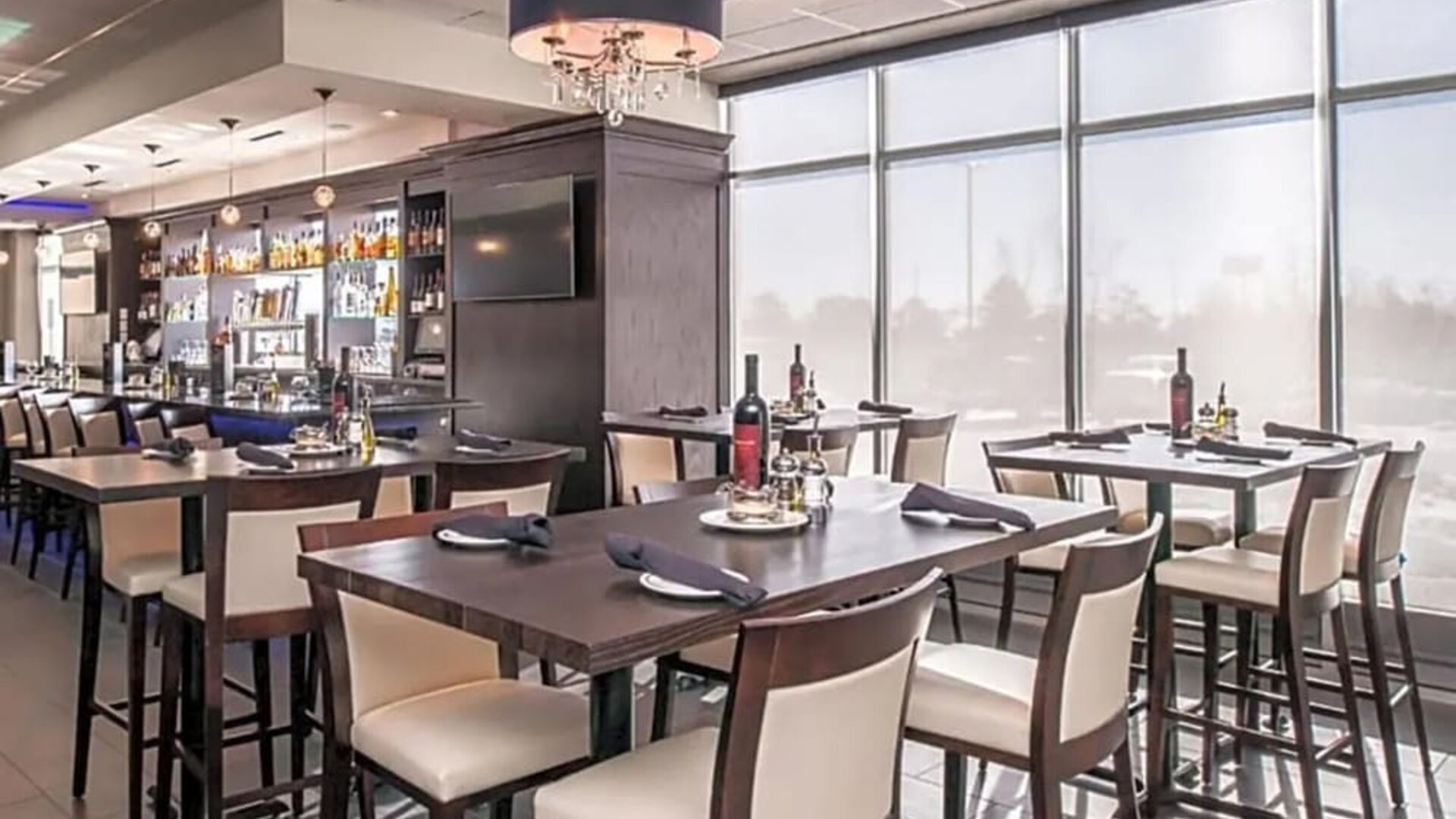 Restaurant dining area with tables, chairs, bar counter, and large windows at Holiday Inn & Suites Farmington Hills - Detroit NW By IHG.