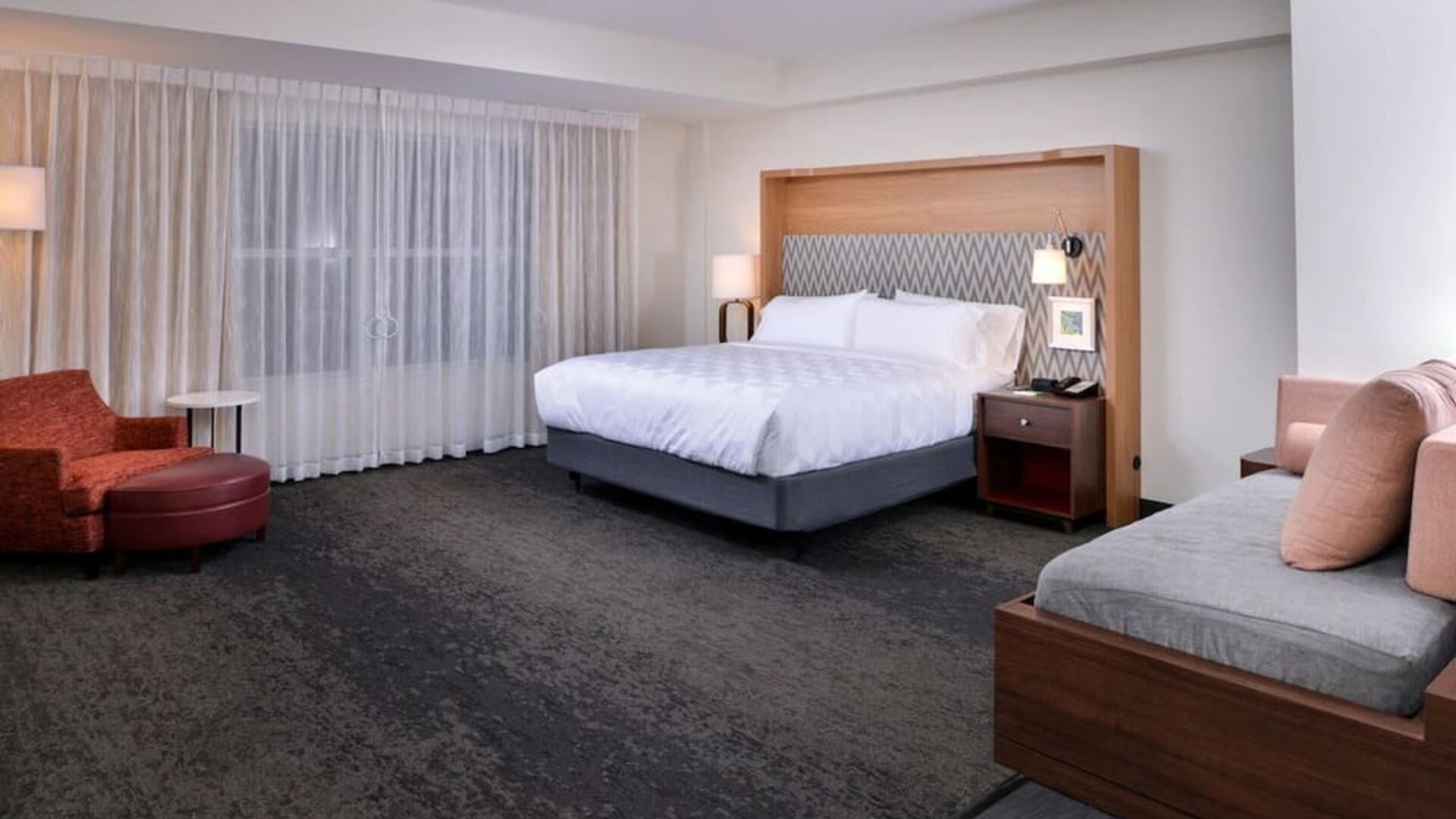 Large suite with king bed, sofa seating, and wide window with sheer curtains at Holiday Inn & Suites Farmington Hills - Detroit NW By IHG.