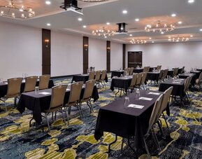 Meeting room with rows of tables, chairs, chandeliers, and patterned carpet at Holiday Inn & Suites Farmington Hills - Detroit NW By IHG.