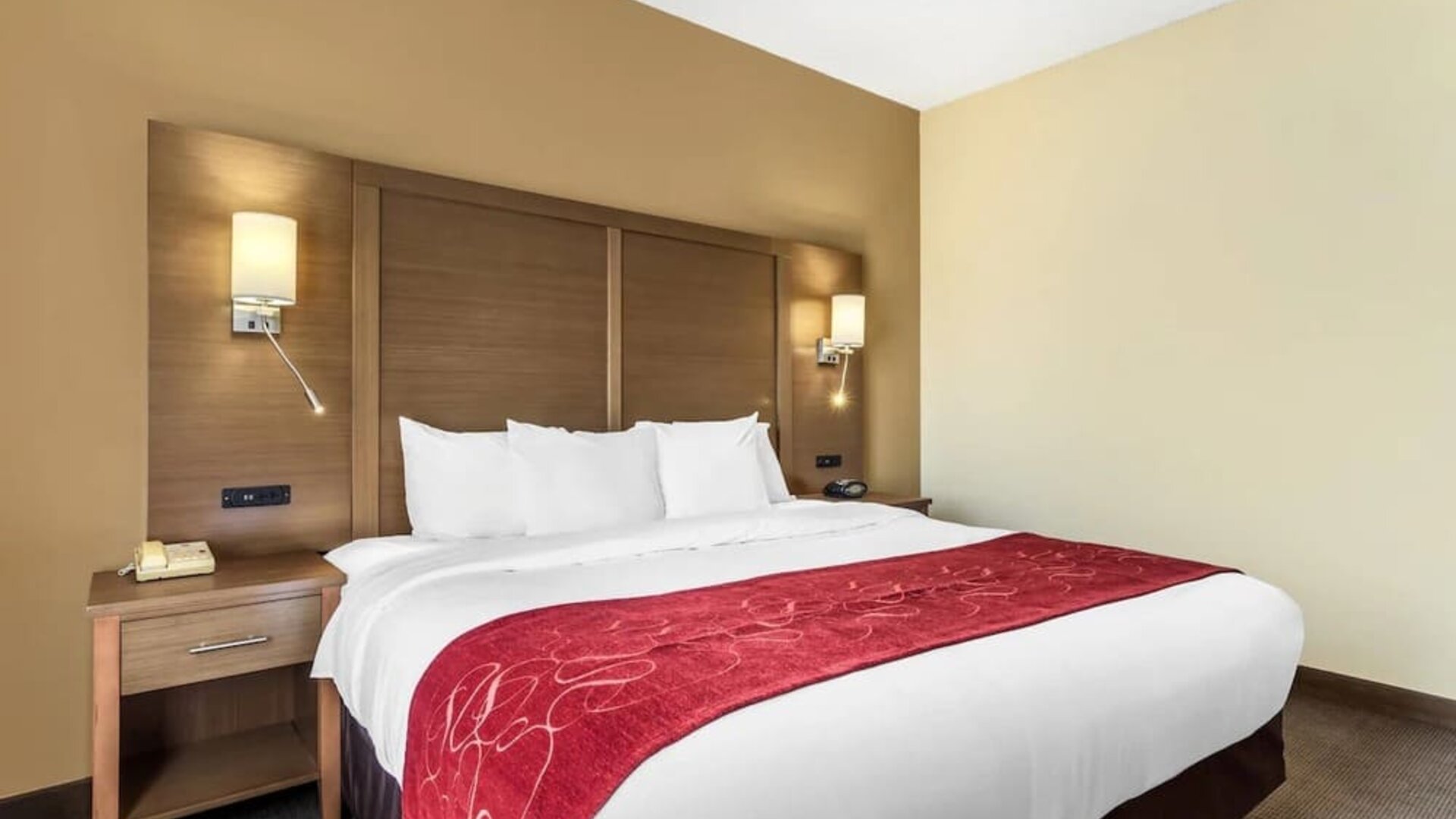 Bed with red runner, wooden headboard, bedside tables, and wall-mounted lights at Comfort Suites Bush Intercontinental Airport.