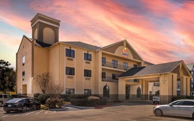 Exterior of hotel building with covered entrance, parked cars, and sunset sky at Comfort Suites Bush Intercontinental Airport.