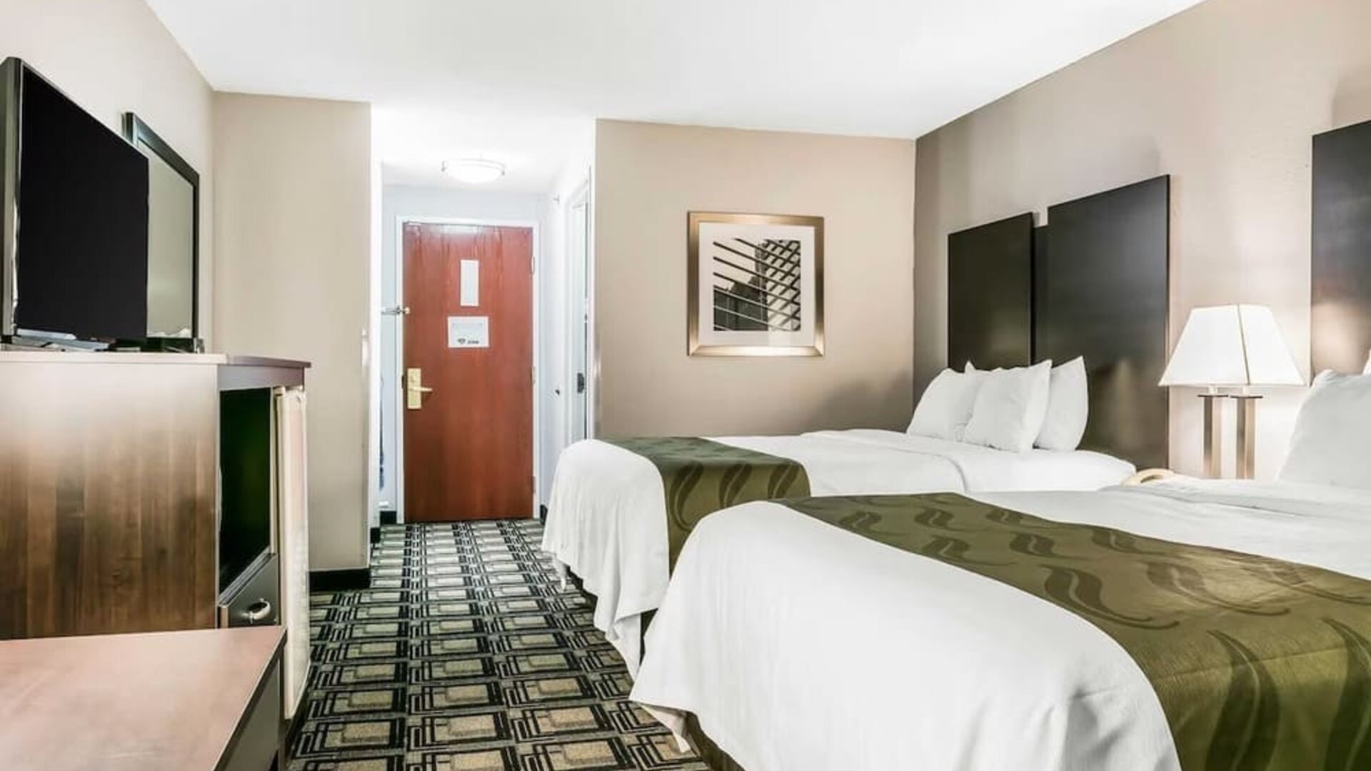 Double room with two beds, bedside table, lamp, and patterned carpet leading to entry door at Suburban Studios Mason Hwy-42.