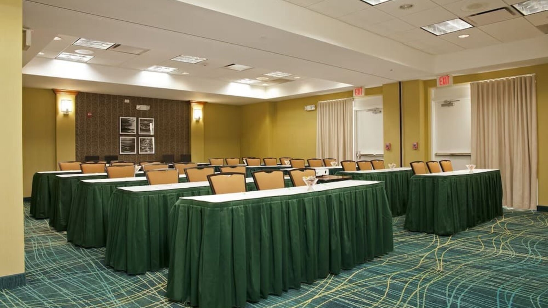 Meeting room with rows of tables draped in green cloths, padded chairs, patterned carpet, and exit doors at Marriott SpringHill Suites Naperville/Warrenville.