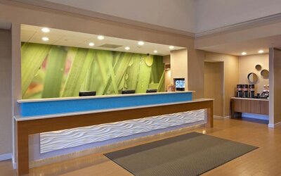 Front desk area with textured counter, green nature mural backdrop, and beverage station to the side at Marriott SpringHill Suites Naperville/Warrenville.