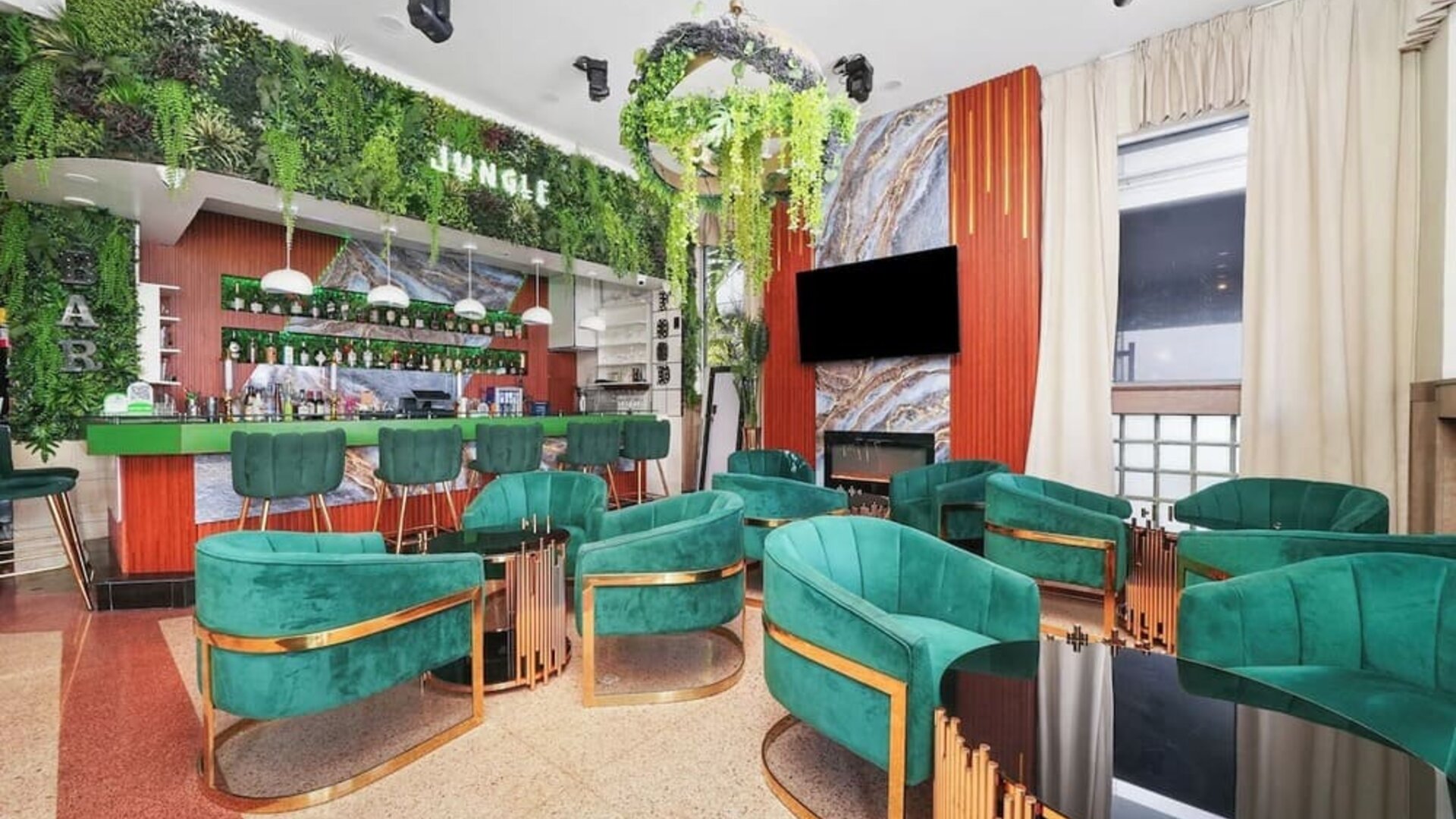 Stylish bar lounge with green velvet chairs, marble accents, hanging plants, and illuminated back bar at Chesterfield Hotel & Suites.
