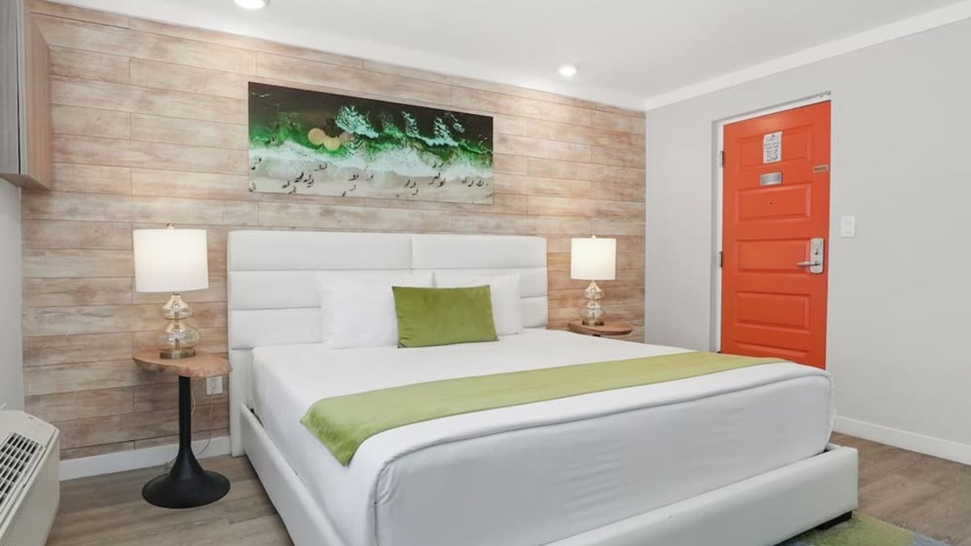 Queen bedroom with upholstered headboard, bedside lamps, artwork, and bright orange entry door at Chesterfield Hotel & Suites.
