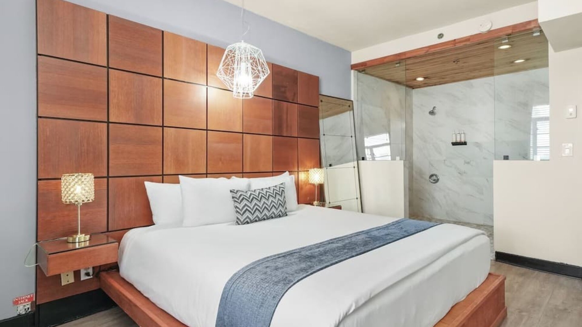 King bedroom with wood-paneled headboard wall, glass-partition shower, and modern lighting at Chesterfield Hotel & Suites.