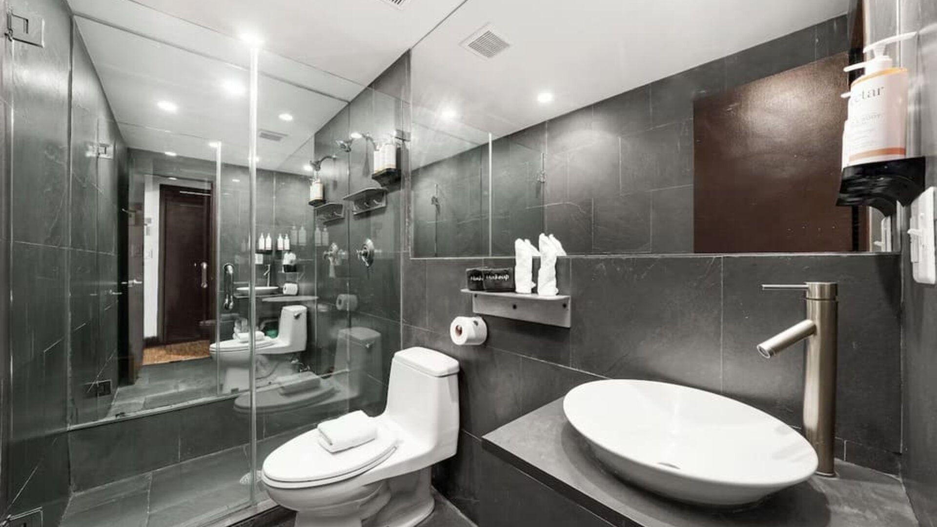 Modern bathroom with dark tile walls, glass shower enclosure, vessel sink, and wall-mounted fixtures at Hotel Chelsea, A South Beach Group Hotel.