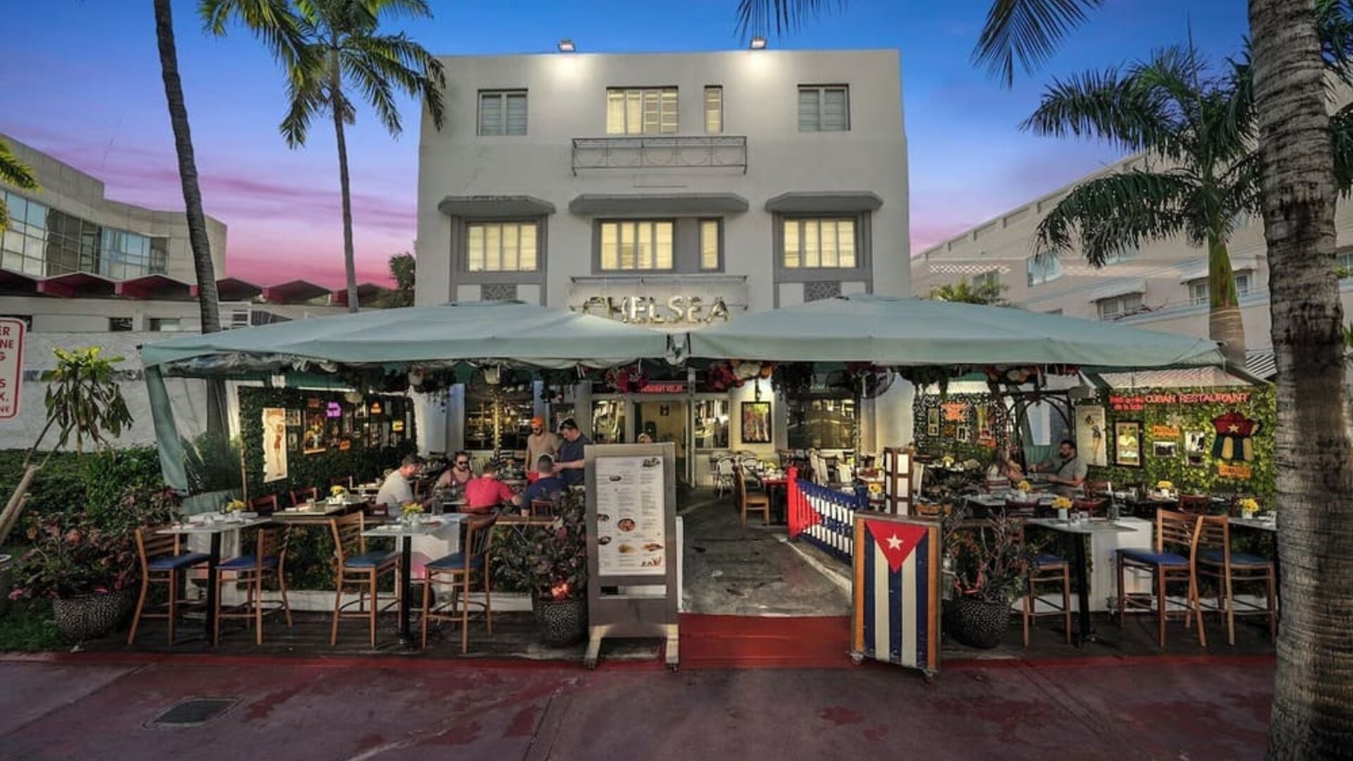 Hotel exterior at dusk with outdoor dining area, palm trees, and lit façade along a lively street at Hotel Chelsea, A South Beach Group Hotel.