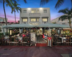 Hotel exterior at dusk with outdoor dining area, palm trees, and lit façade along a lively street at Hotel Chelsea, A South Beach Group Hotel.