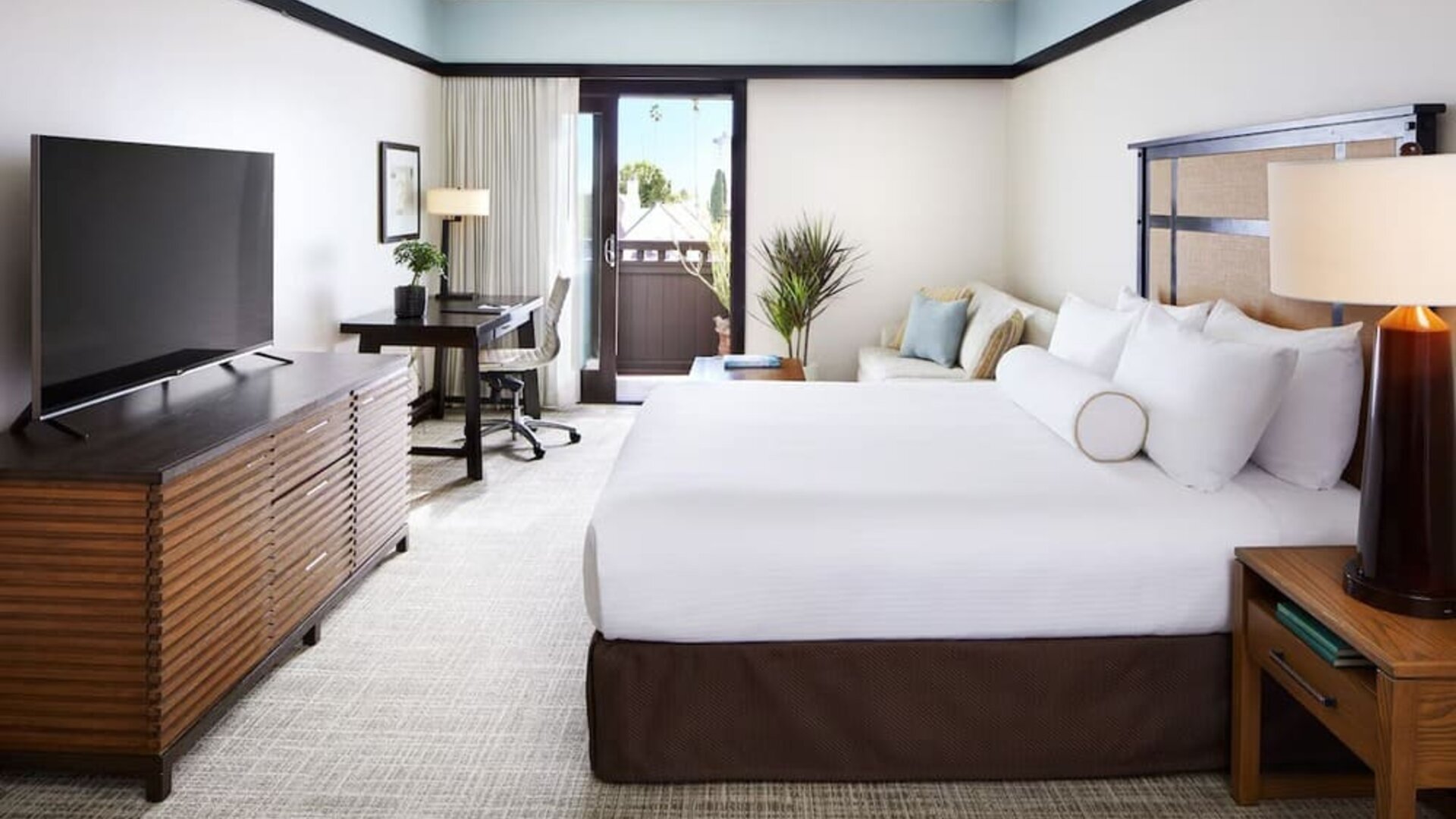 Bright guest room with king bed, desk, TV console, and balcony access with outdoor seating at The Ambrose Hotel Santa Monica.